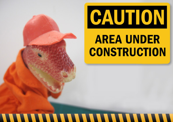 Photo of a t-rex in orange and cap overalls behind a sign that says "Caution, area under construction"